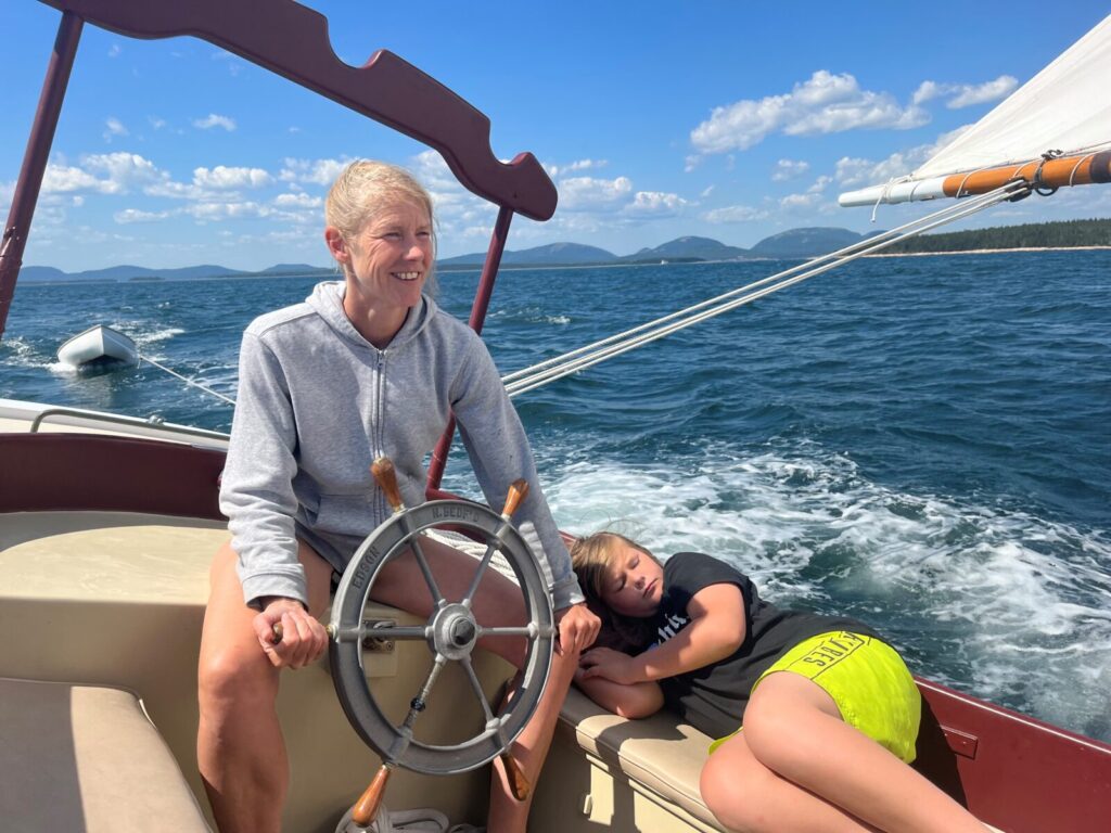 Person steering a sailboat on open water, with a child resting beside them. Sunny day with mountains in the background.