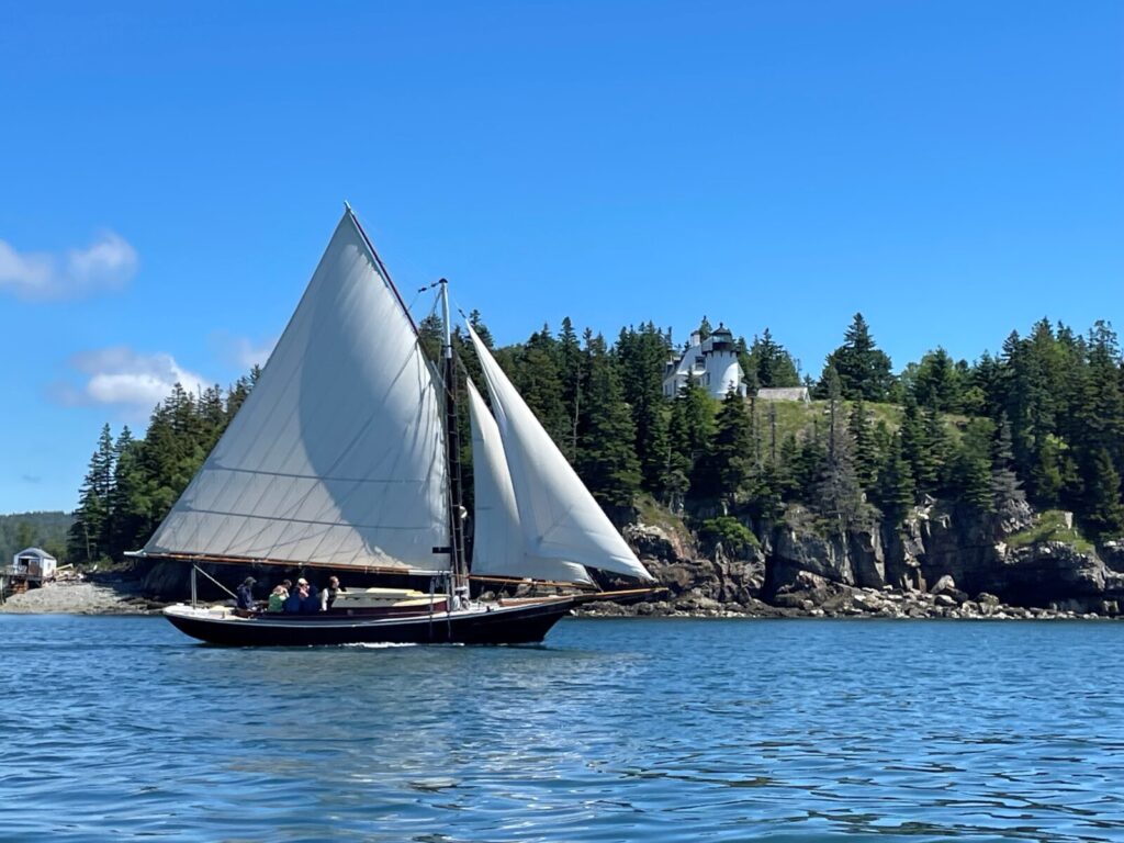 A sailboat with white sails glides on blue water near a rocky shore with trees and a distant lighthouse under blue sky.